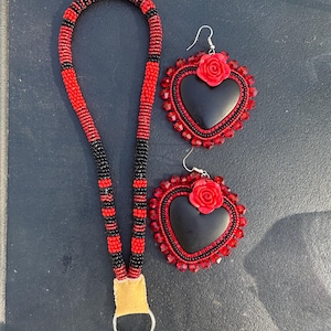 Könnte beinhalten: Ein Perlen-Lanyard mit abwechselnden roten und schwarzen Segmenten, einem beigen Lederverbinder und einem silbernen Metallring, dazu zwei herzförmige Ohrringe mit schwarzer Mitte, roter Rosenapplikation und rotem Kristallperlenrand.
