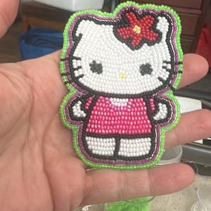 May include: A beaded patch featuring the character Hello Kitty, rendered in white, pink, black, and red beads. The character wears a pink dress and a red flower accessory, outlined with a vibrant lime green beaded border on a dark backing.
