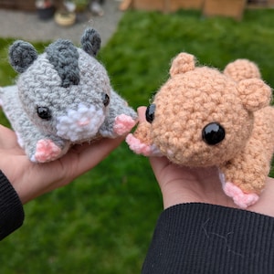 Puede incluir: Dos juguetes de animales hechos a mano con ganchillo. Uno es gris con una raya gris oscuro y detalles en rosa. El otro es de color canela con detalles en rosa. Ambos tienen ojos negros y se sostienen en manos abiertas.