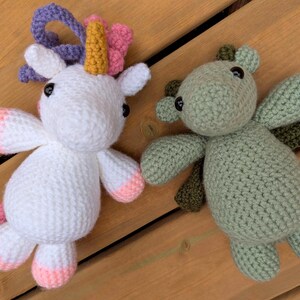 Puede incluir: Dos animales de peluche de ganchillo hechos a mano: un unicornio blanco con detalles rosas y amarillos, y un dragón verde con alas verde oscuro. Ambos tienen ojos de botón negros, sobre un fondo de madera.