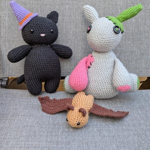 May include: Handmade crochet toys: a black cat with a purple and orange witch's hat, a grey bunny with pink and green details, and a tan bat with brown wings. The toys have button eyes.