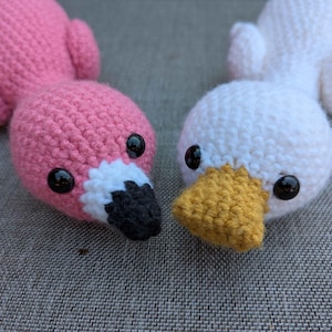 May include: Two crocheted bird toys. One is pink with a black and white beak, and the other is white with a yellow beak. Both have black button eyes and are lying on a grey surface.