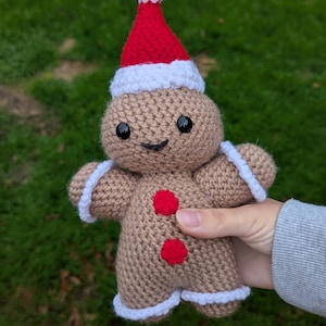 May include: A crocheted gingerbread man doll wearing a red and white Santa hat. The doll has black eyes, a smiling mouth, and two red button accents. White trim outlines the arms and legs. The doll is approximately 15 cm tall.