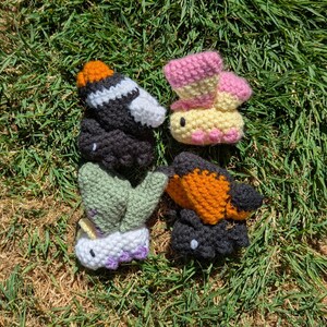 May include: A collection of four small, handmade crochet plush toys shaped like insects. The set includes a black and white bee with an orange top, a pink and yellow butterfly, a green and white caterpillar, and a black and orange butterfly.