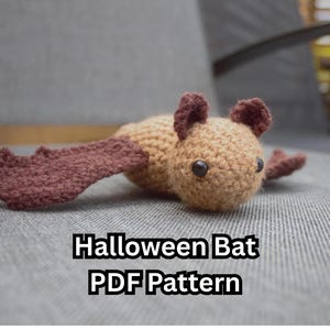 May include: A handmade crochet bat in shades of brown and tan, with dark brown wings and ears. The bat has black button eyes and is lying on a gray surface. Text on the image reads "Halloween Bat PDF Pattern."