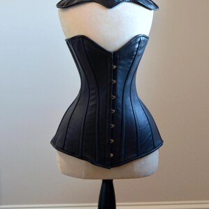 May include: Black leather corset with a matching collar, displayed on a mannequin. The corset features vertical seams and a row of metal clasps down the front. The collar is adorned with silver studs.