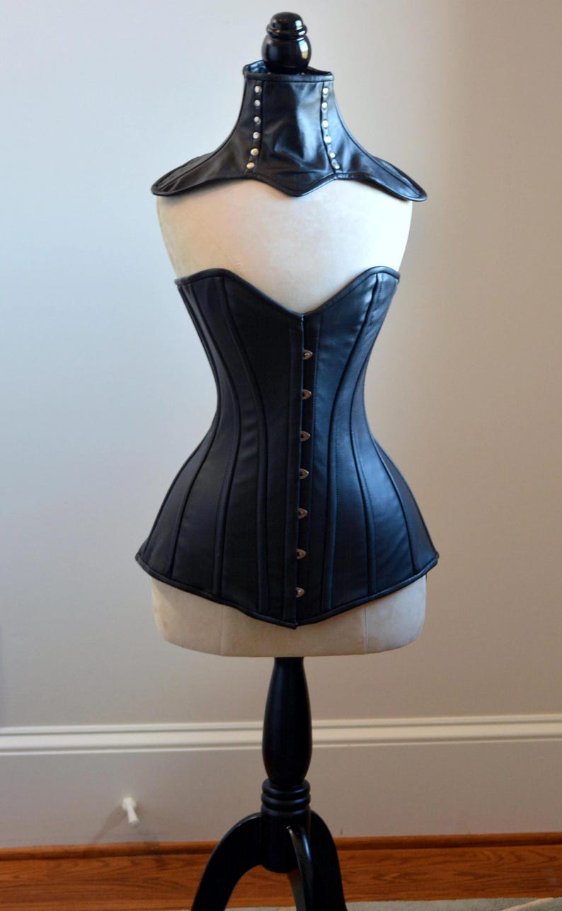 May include: Black leather corset with a matching collar, displayed on a mannequin. The corset features vertical seams and a row of metal clasps down the front. The collar is adorned with silver studs.