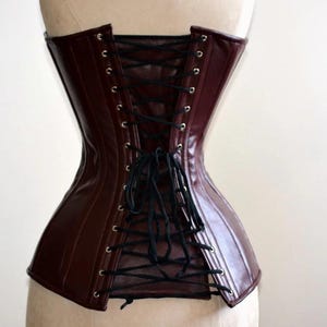 May include: A burgundy corset with black lacing. The corset is made of a shiny material and has a structured design. The back features a lace-up closure with black cording. The corset is displayed on a mannequin.