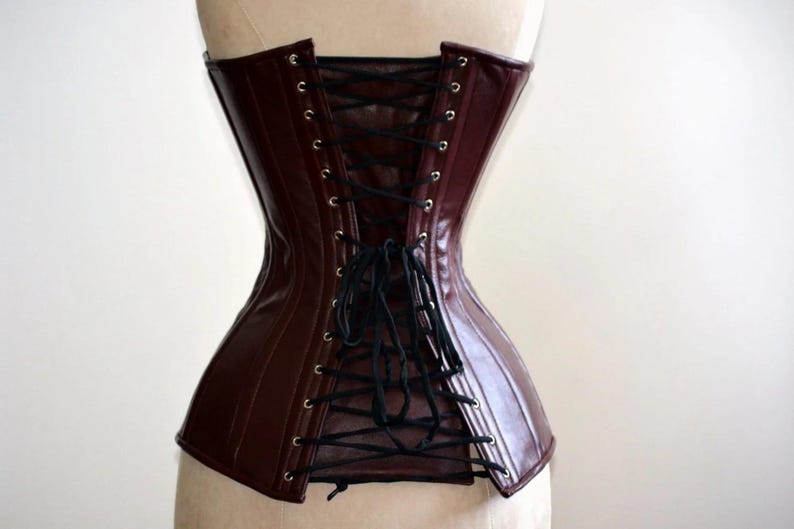May include: A burgundy corset with black lacing. The corset is made of a shiny material and has a structured design. The back features a lace-up closure with black cording. The corset is displayed on a mannequin.