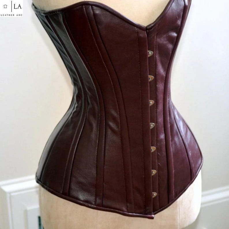May include: A burgundy leather corset with a structured silhouette. The bodice features vertical seams, a curved neckline, and a series of metal clasps down the front. The corset is displayed on a mannequin.