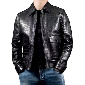 Puede incluir: Una chaqueta negra con un patrón de piel de cocodrilo brillante y texturizado. La prenda incluye un cuello estándar, un cierre de cremallera frontal completo y dos bolsillos rectangulares en el pecho con solapas abotonadas. Las mangas tienen puños abotonados.