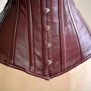 May include: Close-up of a burgundy leather corset. The corset features vertical panels, a pointed bottom edge, and silver-colored clasps. The garment is displayed on a mannequin, showcasing its shape and design.