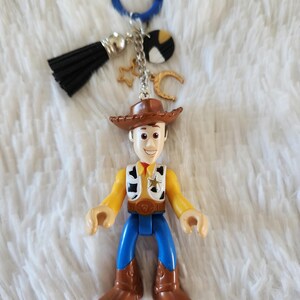 May include: A plastic toy keychain featuring Woody from Toy Story. Woody is wearing his signature cowboy hat, vest, and jeans. The keychain also includes a blue ring, a black tassel, and gold star and crescent moon charms.