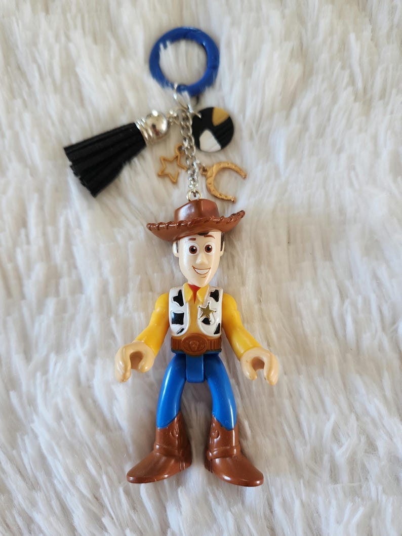 May include: A plastic toy keychain featuring Woody from Toy Story. Woody is wearing his signature cowboy hat, vest, and jeans. The keychain also includes a blue ring, a black tassel, and gold star and crescent moon charms.