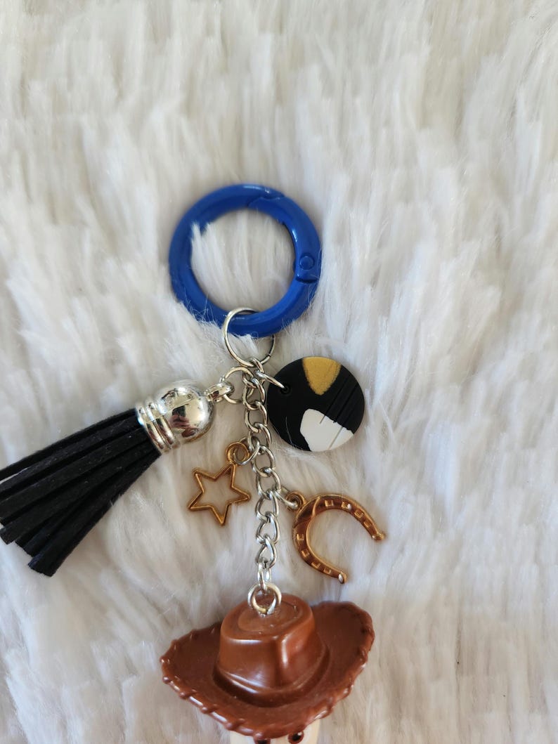 May include: A brown cowboy hat charm with a gold horseshoe, a gold star, and a black tassel attached to a silver chain. The charm is attached to a blue plastic ring.