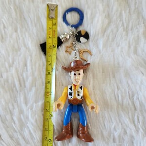 May include: A plastic toy figurine of Woody from Toy Story, wearing his signature cowboy hat, vest, and jeans. The figurine is attached to a keychain with a blue ring, a black tassel, and silver charms including a horseshoe, a star, and a round charm with black and white spots.