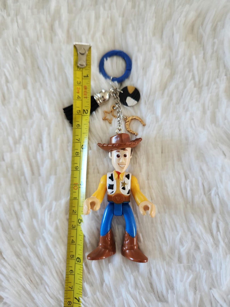 May include: A plastic toy figurine of Woody from Toy Story, wearing his signature cowboy hat, vest, and jeans. The figurine is attached to a keychain with a blue ring, a black tassel, and silver charms including a horseshoe, a star, and a round charm with black and white spots.