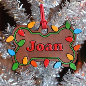 Könnte beinhalten: Ein brauner, knochenförmiger Weihnachtsschmuck mit dem Namen "Joan" in roten Buchstaben. Der Schmuck ist mit bunten Weihnachtslichtern verziert und hängt an einem roten Band. Der Schmuck wird an einem silbernen Lametta-Weihnachtsbaum präsentiert.
