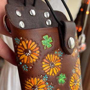 May include: A brown leather phone case with a floral design. The case has a strap and a snap closure. The flowers are painted in shades of orange, yellow, and white. There are also green shamrocks and a bee painted on the case.