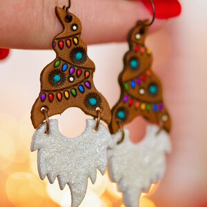 May include: A pair of dangle earrings featuring a brown leather gnome with a white glitter beard. The gnome's hat is decorated with colorful lights and a white pom-pom.