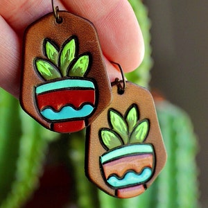 May include: Handmade leather earrings featuring a painted succulent design. The earrings have a brown background with a red, turquoise, and green succulent plant design. The earrings are held up by a person's fingers.