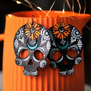 May include: A pair of black sugar skull earrings with white, orange, and turquoise details. The earrings are hanging from a light orange pitcher.