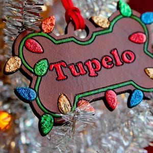 Könnte beinhalten: Ein brauner, knochenförmiger Weihnachtsschmuck mit dem Namen "Tupelo" in roter Schrift. Der Schmuck ist grün umrandet und mit bunten, glitzernden Lichtern verziert. Ein rotes Band ist zum Aufhängen befestigt.
