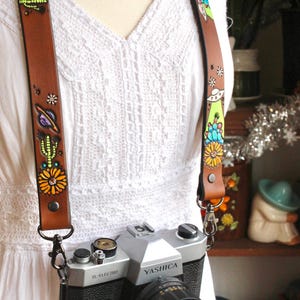 May include: A brown leather camera strap with colorful painted designs of a cactus, flowers, planets, and an alien. The strap is attached to a silver Yashica camera. The strap is worn over a white dress.