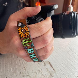 May include: A brown leather camera wrist strap with a hand-painted sunflower design in orange, yellow, and white. The strap also features the word "BKR" in turquoise letters and a green vine design.