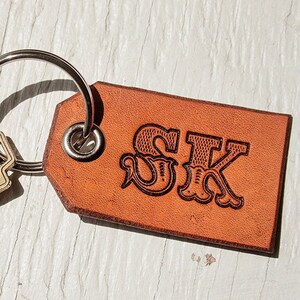 Pode incluir: Um chaveiro de couro castanho com as iniciais "SK" gravadas em uma fonte decorativa. O chaveiro est&aacute; preso a um chaveiro prateado com uma &uacute;nica chave de lat&atilde;o.