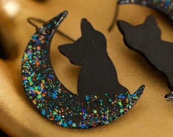 May include: A pair of black cat silhouette earrings with a crescent moon design. The earrings are made of black polymer clay and have a glittery finish with blue, green, and orange glitter.