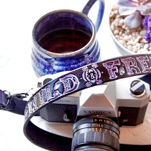 May include: A black leather camera strap with the words "WILD & FREE" embossed in purple. The strap is attached to a silver camera with a black lens.