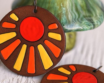 Pode incluir: Um par de brincos de couro marrom com um design de sol. O sol tem um centro vermelho e raios amarelos e laranja.
