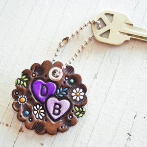 May include: A brown leather keychain with two purple hearts, one with the letter "D" and the other with the letter "B". The hearts are surrounded by colorful flowers and leaves. The keychain is attached to a silver key ring and a silver key.