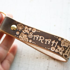 May include: A brown leather keychain with a gold key ring and the name "SARAH" embossed in the leather. The keychain is decorated with floral and butterfly designs.