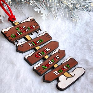 Könnte beinhalten: Ein hölzerner Weihnachtsschmuck in Form von Straßenschildern. Die braunen Schilder haben weiße Schneebesonderheiten und die Namen "The Griswolds", "Ellen", "Clark", "Audrey" und "Rusty" in verschiedenen Farben. Ein rotes Band ist zum Aufhängen befestigt.