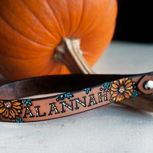 May include: A brown leather keychain with a floral design and the name "Alannah" written in white lettering. The keychain has a silver ring attached to it.