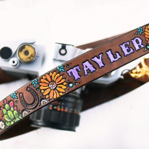 May include: Brown leather camera strap with a purple inscription that says "TAYLOR" and colorful floral and cactus designs. The strap also features a horseshoe design.