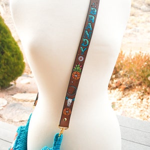 May include: A brown leather bag strap with a colourful design featuring flowers, cacti, a skull, and the name "BRANDY" written in blue and green. The strap is attached to a turquoise crocheted bag.