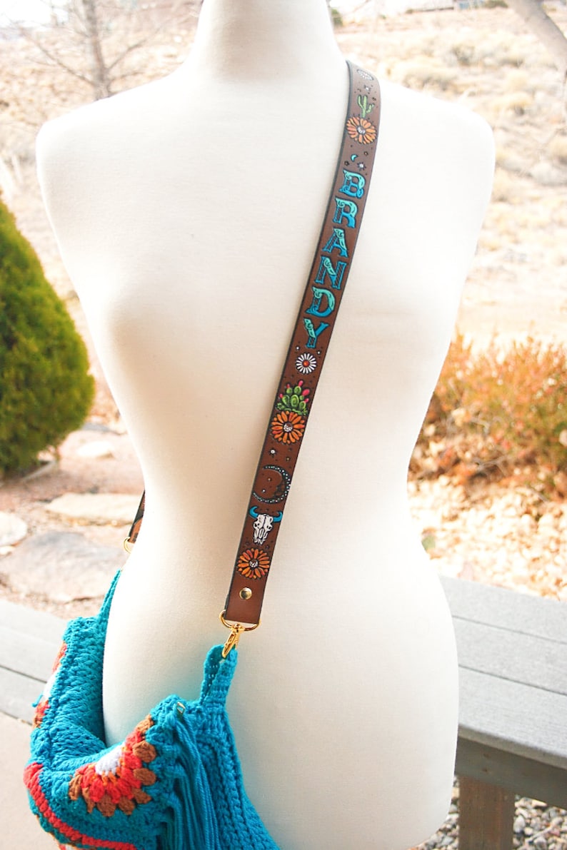 May include: A brown leather bag strap with a colourful design featuring flowers, cacti, a skull, and the name "BRANDY" written in blue and green. The strap is attached to a turquoise crocheted bag.