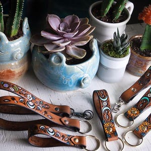 May include: An assortment of brown leather keychains with floral and crescent moon designs. The keychains are displayed with various potted succulents and cacti in ceramic pots, creating a rustic, bohemian aesthetic.