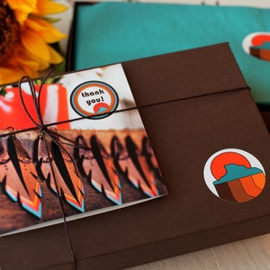 May include: A brown gift box with a thank you card and a sticker featuring a sunset graphic. The box is tied with twine and has a sticker that says "thank you!" The card shows a photograph of colourful feathers.