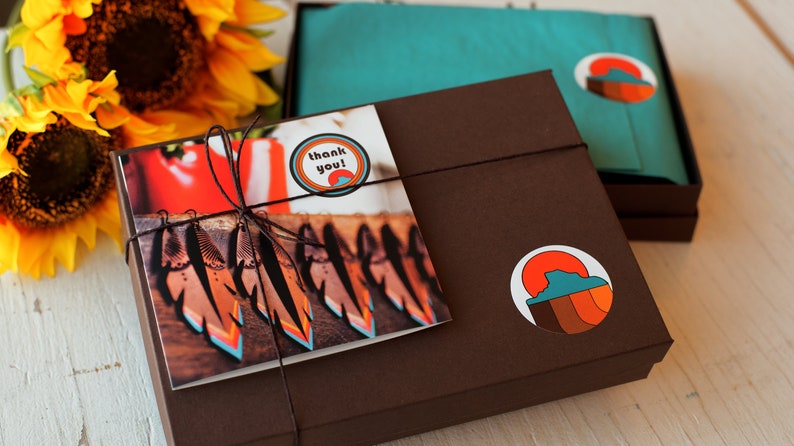 May include: A brown gift box with a thank you card and a sticker featuring a sunset graphic. The box is tied with twine and has a sticker that says "thank you!" The card shows a photograph of colourful feathers.