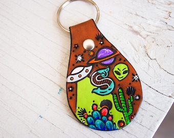 May include: A brown leather keychain featuring a green, purple and white alien, spaceship and cactus design.