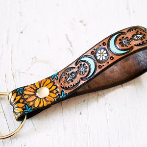 May include: A brown leather keychain with a floral and moon design. The keychain has a gold metal ring.