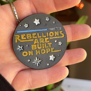 以下が含まれることがあります： 「REBELLIONS ARE BUILT ON HOPE」の文字が金色で書かれた、丸い黒色の装飾品。青いライトセーバー、白い星、雪の結晶のデザインが含まれています。手で持っています。
