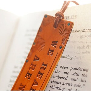 May include: A brown leather bookmark with the text "WE REAL ARE N" embossed on it. The bookmark has a blue tassel and a decorative design.