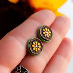 May include: A pair of green and brown stud earrings with a yellow flower design. The earrings are on a person's hand.