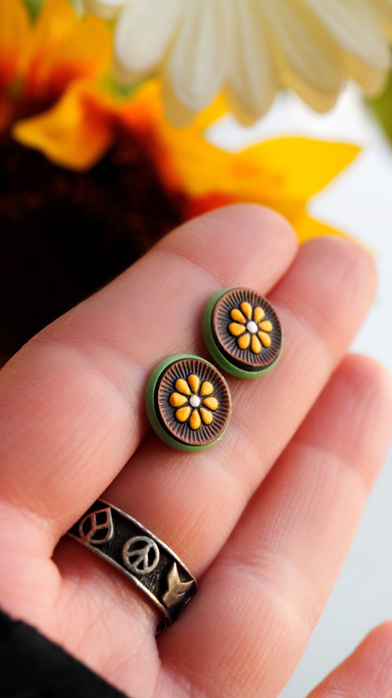 May include: A pair of green and brown stud earrings with a yellow flower design. The earrings are on a person's hand.