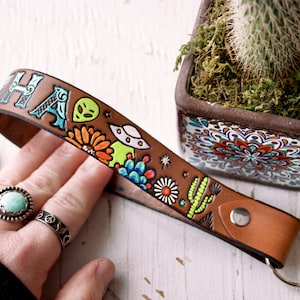 May include: A brown leather keychain with a colorful design featuring the word "HA" and various illustrations, including cacti, flowers, and a UFO. The keychain has a silver ring.